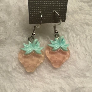 Jordgubbsörhängen i pastell - Säljer ett par söta örhängen med jordgubbsformade hängen i ljusrosa och mintgrönt. Perfekta för att ge en lekfull touch till din stil. Hängena är i plast och öronkrokarna i sterling silver 