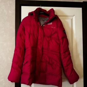 Rosa dunjacka med huva från Röhnisch - Säljer en rosa dunjacka från Röhnisch med huva, dragkedja framtill och dragsko i midjan. Jackan har långa ärmar, två fickor med dragkedja och quiltad design. Perfekt för kyliga dagar och håller dig varm hela säsongen.