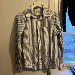 Randig skjorta från Abercrombie & Fitch - Snygg skjorta med blå och vita ränder från Abercrombie & Fitch. Klassisk krage, bröstficka med broderad älg och knäppning framtill. Perfekt för en stilren look.