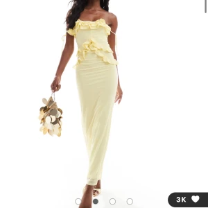 Ljusgul långklänning med volangdetaljer - Supersnygg ljusgul långklänning med volanger och axelbandslös design. Klänningen har en figurnära passform och knytband på axeln för extra detalj. Perfekt för sommarens speciella tillfällen.