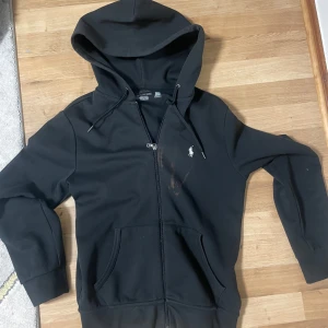 Svart hoodie med dragkedja från Polo Ralph Lauren - Svart hoodie med dragkedja och huva från Polo Ralph Lauren. Klassisk design med vit broderad logga på bröstet och två fickor framtill. Justerbar huva med snörning och ribbade muddar vid ärmslut och nederkant.