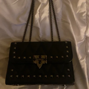 Svart axelväska med nitar och kedjerem - Säljer en svart axelväska med quiltad design och silverfärgade nitar på framsidan. Väskan har en snygg kedjerem och ett metallspänne framtill. Perfekt för dig som vill ha en edgy accessoar till din outfit.