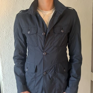 Mörkblå fieldjacka  - Snygg mörkblå fieldjacka med fyra framfickor och knappar. Jackan har krage, axeldetaljer och är långärmad. Passformen är normal och materialet känns som syntet eller polyester. Perfekt för vår och höst. Storlek M men sitter mer som S. 