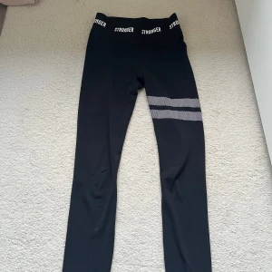 Svarta leggings från Stronger med vita prickiga ränder - Säljer ett par svarta leggings från Stronger i storlek XS. De har två vita prickiga ränder på ena benet och elastisk midja med logga. Tillverkade i polyester och elastan, perfekta för träning.