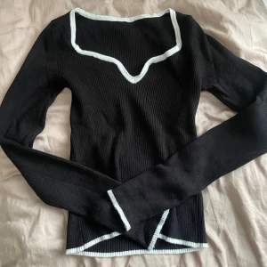 Svart ribbad långärmad topp med vit kant - Säljer en svart ribbad långärmad topp med vit kant längs halsringning, ärmslut och nederkant. Toppen har en unik v-ringad design och sitter snyggt på kroppen. Perfekt att styla med jeans eller kjol!