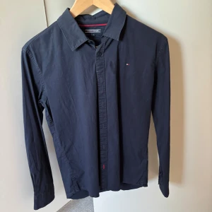 Mörkblå skjorta från Tommy Hilfiger - Snygg mörkblå skjorta från Tommy Hilfiger med klassisk krage och diskret logga på bröstet. Skjortan har långa ärmar och knäppning framtill. Perfekt för en stilren look.