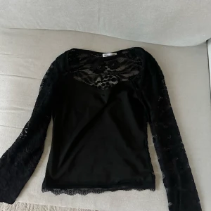 Venderbys Svart spetsblus med lång ärm - Säljer en svart blus med långa ärmar i genombruten spets och fina spetsdetaljer vid ärmslut och nederkant. Toppen har en croppad passform och v-ringning framtill. Perfekt för dig som gillar detaljer och en trendig look. OBS!!! Litet hål vid ärmen, syns på bild 3. Strl s/m. Använd 1 gång. 