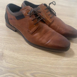 Bruna skinnloafers med snörning - Säljer ett par bruna loafers i skinn med snörning och diskret textur. Skorna har en klassisk design med spetsig tå och svarta detaljer vid hälen. Perfekta för en dressad look.