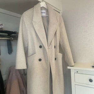 Beige kappa från Bershka - Säljer en stilren beige kappa från Bershka med dubbelknäppning och bred krage. Jackan har långa ärmar och två fickor framtill. Perfekt för dig som vill ha en klassisk och tidlös look.