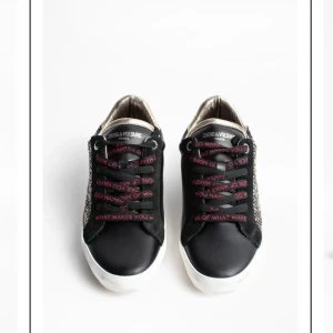 Svarta sneakers från Zadig & Voltaire med nitar - Säljer ett par svarta sneakers från Zadig & Voltaire med metallicguld häl, svarta snören med röd text och coola silverfärgade nitar på sidorna. Skorna har vit sula och är aldrig använda trots att de är slitna (del av stylen). Storlek 38 och är snygga sommarsneakers 🌸
