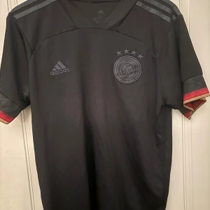Svart Tyskland fotbollströja från Adidas - Säljer en svart Tyskland fotbollströja från Adidas med fyra stjärnor och förbundets emblem på bröstet. Tröjan har korta ärmar med röd och gul detalj vid ärmslut samt Adidas-logga på bröstet och axelränder.