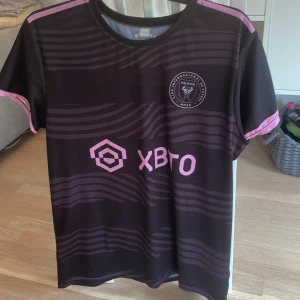 Inter Miami CF Messi 10 fotbollströja - Säljer en svart Inter Miami CF fotbollströja med rosa detaljer och Messi 10 på ryggen. Tröjan har korta ärmar, klubbmärke på bröstet och är tillverkad i 100% polyester. Perfekt för fans av Messi och MLS.