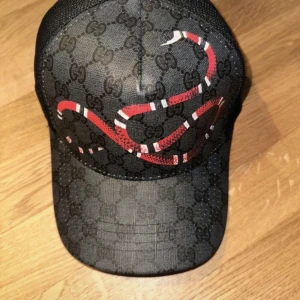 Gucci Keps - Snygg svart keps från Gucci med klassiskt GG-mönster och en röd, vit och svart orm på framsidan. Kepsen har nät på baksidan och justerbar passform. Perfekt för dig som vill sticka ut med en exklusiv accessoar.