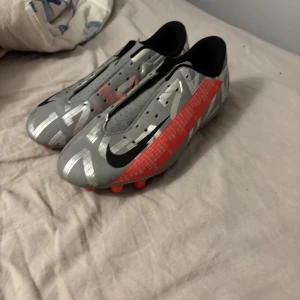 Nike Mercurial fotbollsskor i silver och rött - Säljer ett par Nike Mercurial fotbollsskor i silver med röda och svarta detaljer. Skorna har snörning, rund tå och platt sula med röda dobbar. Perfekta för dig som vill sticka ut på planen med snygg design och bra grepp.
