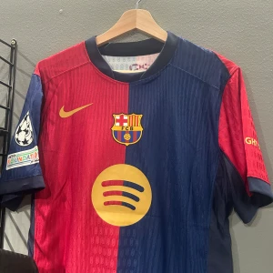 FC Barcelona matchtröja med Lamine Yamal 19 - Säljer en officiell FC Barcelona fotbollströja med Lamine Yamal och nummer 19 på ryggen. Tröjan har klassiska blå och röda färger framtill, svarta detaljer baktill, samt klubbmärke och sponsorlogga på bröstet. Kortärmad modell med Champions League-märke på ärmen.