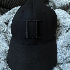 Svart keps från Les Deux - Snygg svart keps från Les Deux med broderad logga framtill och justerbar snapback baktill. På sidan finns små broderade ränder i vitt och rött. Perfekt accessoar för en stilren look.  Använd endast 3-4 gånger