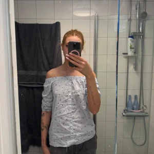 Offshoulder randig skjorta från Atmosphere - Säljer en ljusblå och vit randig offshoulder-skjorta från Atmosphere med små blommiga detaljer. Skjortan har knappar framtill och resår upptill för en snygg passform. Perfekt för dig som gillar en avslappnad men trendig stil.