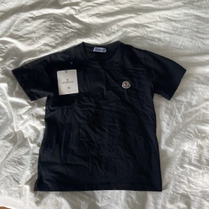 Svart t-shirt från Moncler - Säljer en svart t-shirt från Moncler med klassisk rund hals och broderad logotyp på bröstet. T-shirten är i mjuk bomull och har en normal passform. Perfekt för dig som gillar stilrena och exklusiva plagg.