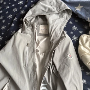 Ljusgrå vindjacka Moncler - Säljer en stilren ljusgrå vindjacka från Moncler med huva och dragkedja framtill. Jackan har en bröstficka med dragkedja, Moncler-logga på ärmen och diskret tryck på framsidan. Perfekt för blåsiga dagar.