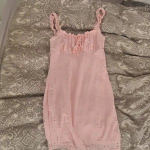 Ljusrosa klänning med volangband från SHEIN - Supersöt ljusrosa klänning från SHEIN med volangprydda axelband och knytning framtill. Klänningen har broderade hålmönster och en feminin, figurnära passform. Perfekt för sommarens alla tillfällen!🎀