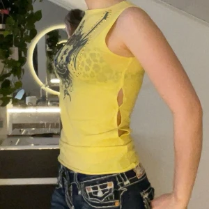 Gul ärmlös topp/linne med tryck och nitar - Snygg gul ärmlös topp med ett stort svart tryck av ett lejon och svärd på bröstet. 