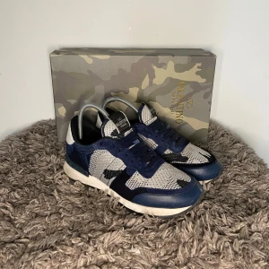 Valentino Rockrunners - Säljer ett par snygga Valentino Garavani sneakers i blått, grått och vitt med kamouflagemönster. Skorna har snörning, rund tå och platt sula. Perfekta för dig som vill sticka ut med stilrena och exklusiva sneakers.
