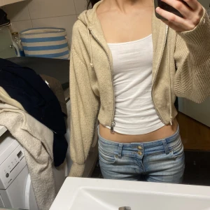 Beige croppad stickad hoodie med dragkedja - Säljer en beige croppad stickad hoodie med dragkedja framtill och huva. Jackan har långa ärmar och ribbade muddar. Perfekt för en avslappnad och trendig look.