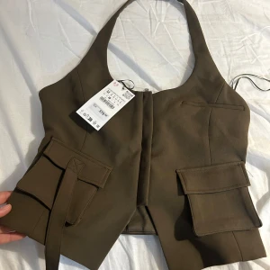 Brun väst med fickor från Zara - Trendig brun väst från Zara med breda axelband och djup urringning. Västen har två stora fickor framtill och stängs med dragkedja i ryggen. Perfekt att styla över en skjorta eller t-shirt för en cool look.