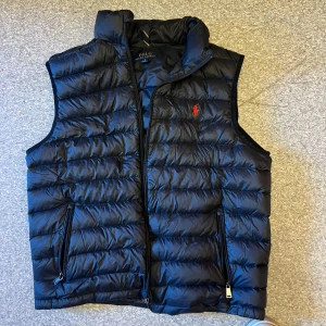 Svart dunväst från Polo Ralph Lauren - Säljer en svart dunväst från Polo Ralph Lauren med klassisk quiltad design och röd broderad logga på bröstet. Västen har dragkedja framtill och två fickor med dragkedja. Perfekt för lager-på-lager under kyliga dagar.