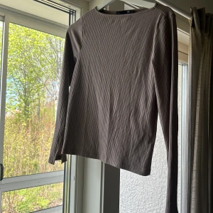 Ribbad långärmad topp i brun - Säljer en stilren, ribbad långärmad topp i brun färg. Toppen har en rund halsringning och är tillverkad i ett mjukt, stretchigt material som sitter bekvämt på kroppen. Perfekt för lager-på-lager eller som den är. Har även en liten slits vid ärmen.