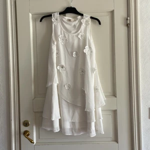 Vit ärmlös klänning med blommor från Aura - Säljer en vit ärmlös klänning från Aura med lager på lager-effekt och dekorativa blommor med pärldetaljer framtill. Klänningen har en lös passform och är perfekt för sommarens tillfällen.