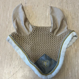 Trinity huva - Snygg beige ridhuva med långa öron och dekorativ kant i vitt och guld. Nätstickad mittdel och tyg vid öronen, perfekt för ridning. Stl full 