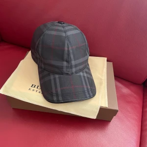 Rutig keps från Burberry - Snygg keps från Burberry i klassiskt rutigt mönster i svart och grått med diskreta röda linjer. Kepsen har böjd skärm och justerbar passform. Perfekt accessoar för en stilren look.