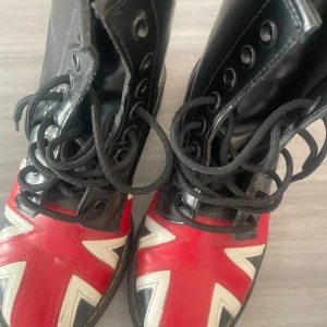Svarta boots med Union Jack-mönster - Säljer ett par svarta boots med snörning och Union Jack-flagga i rött, vitt och blått på tån. Skorna har högre skaft och klassisk rund tå. Perfekt för dig som vill sticka ut med en unik stil!