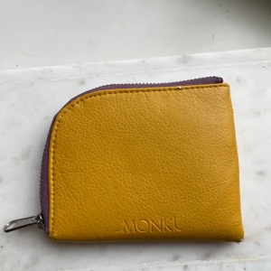 Gul plånbok från Monki - Säljer en liten gul plånbok från Monki med lila dragkedja och insida. Perfekt för kort och mynt, smidig att ha med sig i väskan. Gjord i syntetmaterial och har ett stilrent utseende.