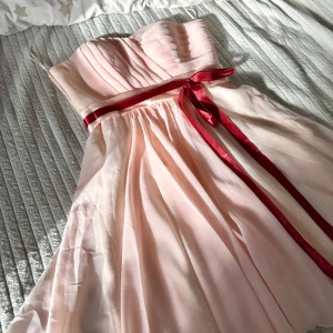 Ljusrosa axelbandslös klänning med röd rosett - Söt ljusrosa axelbandslös klänning med plisserad överdel och röd satinrosett i midjan. Kjolen är lätt och luftig med fint fall. Perfekt för bal eller festliga tillfällen.