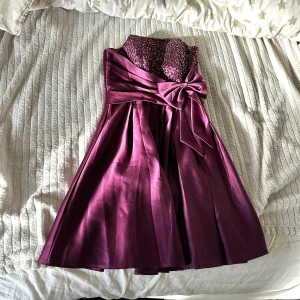 Lila  klänning med paljetter och rosett - Vinröd axelbandslös klänning med glittrig paljettöverdel och stor rosett i midjan. Kjolen är utsvängd och materialet har en satinliknande glans. Perfekt för festliga tillfällen där du vill sticka ut.
