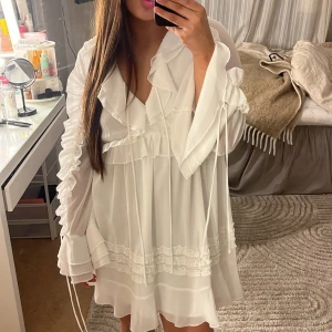 Vit volangklänning med knyt och långa ärmar - Säljer en söt vit klänning med volangdetaljer och knyt framtill. Klänningen har långa, vida ärmar och flera lager volanger nertill. Perfekt för högtider som tex student/ midsommar💗endast andvänd 2 gånger 