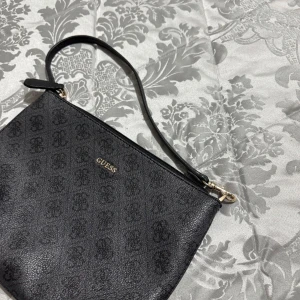 Svart axelväska från Guess - Säljer en stilren svart axelväska från Guess med diskret logomönster och guldfärgade detaljer. Väskan har dragkedja och ett ljust innerfoder med Guess-logga. Perfekt storlek för det viktigaste och enkel att bära över axeln.