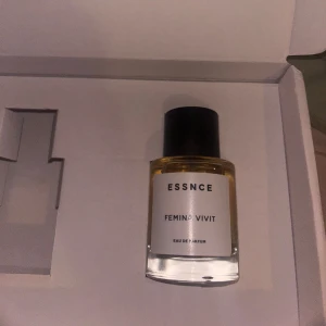 ESSNCE Femina Vivit Eau de Parfum - En stilren parfym från ESSNCE med namnet Femina Vivit. Flaskan är genomskinlig med en vit etikett och svart text, samt ett svart cylinderformat lock. Parfymen har en elegant och minimalistisk design som passar perfekt i badrumshyllan. Bara testad
