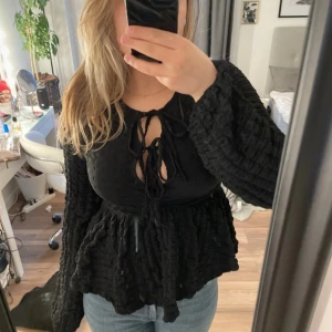 Svart blus med knytning från Gina Tricot - Säljer en svart blus från Gina Tricot med långa, vida ärmar och knytning framtill. Blusen har en strukturerad, lätt rynkad yta och markerad midja. Perfekt för dig som gillar trendiga detaljer och en unik look.