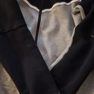 Svart och grå naike tech - Säljer en naike tech p-jacka i svart och grått med vit detalj längs bröstet. Jackan har lång ärm och dragkedja framtill. Perfekt för en avslappnad stil eller till träning.