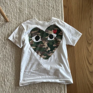 Cdg T-shirt  - Vit t-shirt med stort hjärta i camouflage-mönster och ögon på bröstet. Liten röd broderad hjärtdetalj med ögon uppe till höger på hjärtat. Klassisk rund hals och korta ärmar.