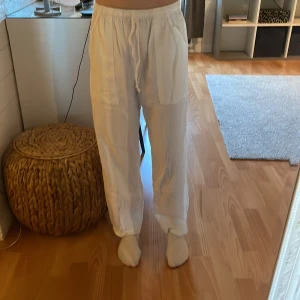 Vita linnebyxor med resårmidja - Säljer ett par vita, luftiga linnebyxor i storlek M med resår i midjan och raka ben. Byxorna är tillverkade i 100% bomull och har en avslappnad passform, perfekta för varma dagar.