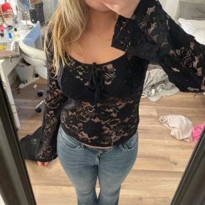 Svart spetsblus från Vera Moda - Säljer en svart, transparent spetsblus från Vera Moda med långa utsvängda ärmar och knytning framtill. Blusen har ett blommigt spetsmönster och är croppad i modellen. Perfekt för dig som vill ha en trendig och snygg topp!