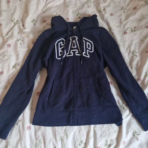 Mörkblå hoodie från GAP - Super snygg mörkblå hoodie från GAP med dragkedja och vit broderad logga framtill. Tröjan har långa ärmar, huva med dragsko och två fickor framtill. Den är tillverkad i bomull och polyesterblandning. Den är nästan aldrig använd!❤️ (pris går att diskutera🤗)