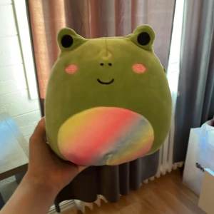 Grön Squishmallows groda med regnbågsfärgad mage - Supermjuk och gosig grön Squishmallows-groda med söta rosa kinder och ett glatt ansikte. Magen är regnbågsfärgad i pastelltoner. Perfekt som kramdjur eller dekoration i rummet.