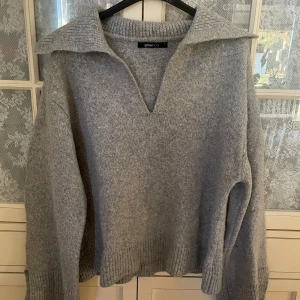 Grå stickad tröja med krage - Mysig grå stickad tröja med bred krage och djup v-ringning. Tröjan har långa ärmar och ribbade muddar vid ärmslut och nederkant. Perfekt för en avslappnad men samtidigt stilren look🤍