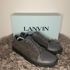 Lanvin Cap Toe - Säljer ett par stilrena grå sneakers från Lanvin med ovandel i mocka och tåparti i skinn. Skorna har snörning och svart platt sula. Perfekta för dig som gillar minimalistisk design och hög kvalitet.