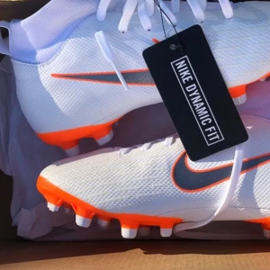 Vita och orangea Nike fotbollsskor - Säljer ett par fräscha vita Nike fotbollsskor med orange detaljer och svart swoosh. Skorna har snörning och platt sula med dobbar, perfekta för gräsplan. Snygg och sportig design med Dynamic Fit. PRIS KAN DISKUTERAS.